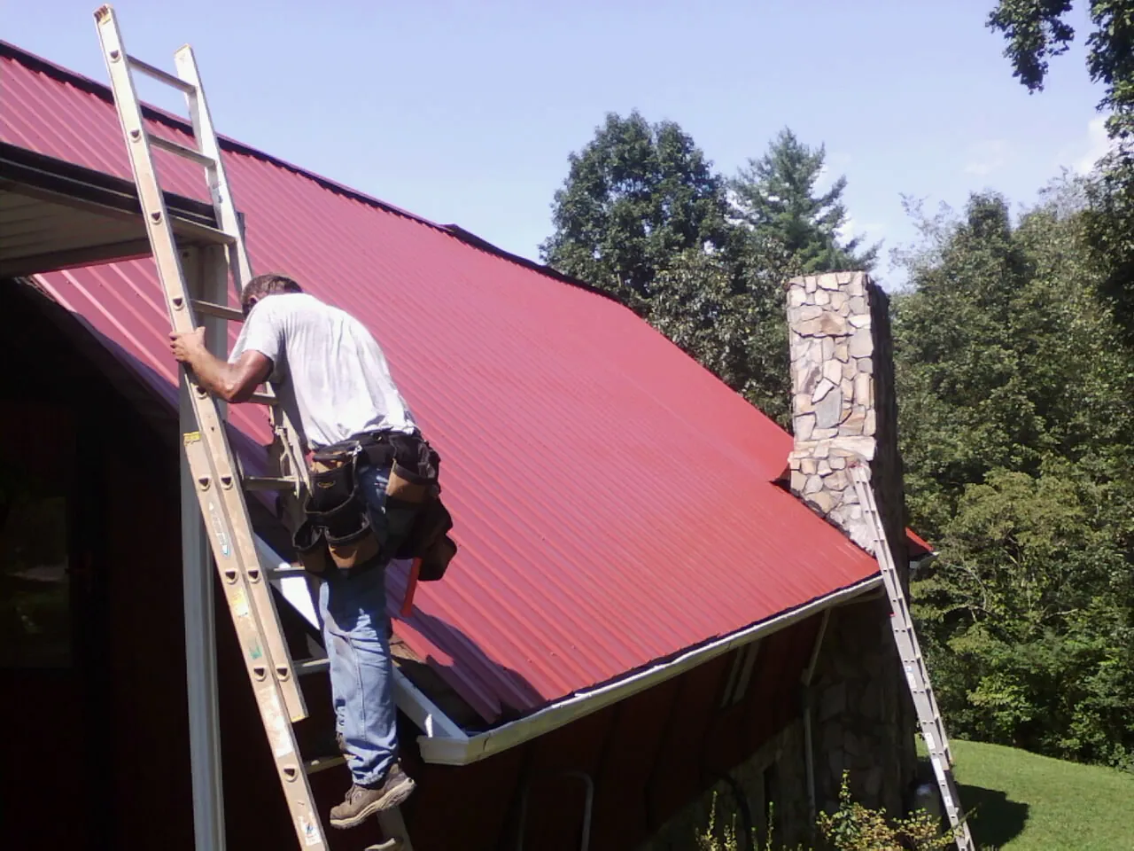 Roofer on a ladder beside a red metal roof and stone chimney — Soffit & Fascia Repair by Roofing Brothers in Rock Island